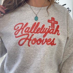 Może przedstawiać: Jasnoszara bluza z napisem "Hallelujah Hooves" w czerwonym skrypcie, wraz z czerwoną grafiką krzyża. Bluza wykonana jest z miękkiego materiału. Widoczny jest srebrny naszyjnik z turkusowym wisiorkiem.