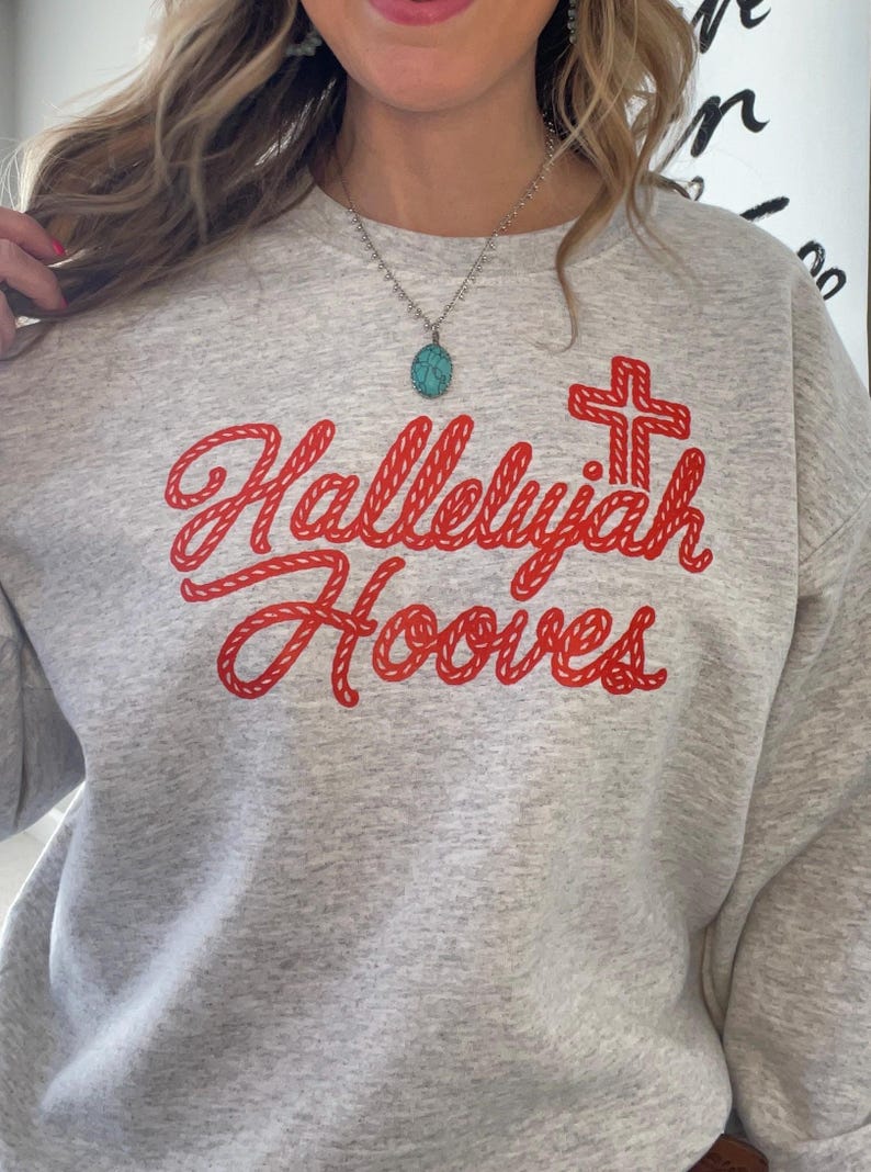 Może przedstawiać: Jasnoszara bluza z napisem "Hallelujah Hooves" w czerwonym skrypcie, wraz z czerwoną grafiką krzyża. Bluza wykonana jest z miękkiego materiału. Widoczny jest srebrny naszyjnik z turkusowym wisiorkiem.