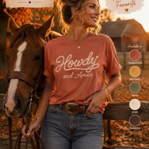 May include: A terracotta-colored short-sleeve t-shirt featuring the text Howdy and Amen in a white rope-style font. The shirt is labeled Comfort Colors 1717 and is displayed alongside a color palette showing terracotta, mustard, moss, ivory, and espresso.