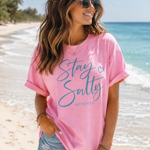 Könnte beinhalten: Ein hellrosa T-Shirt mit kurzen Ärmeln, das auf der Vorderseite den Schriftzug Stay Salty in dunkelblauer Schreibschrift zeigt, ergänzt durch ein kleines Herzsymbol und den Verweis Matthew 5:13 in einer kleineren, serifenlosen Schrift.