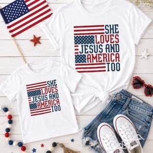 May include: Two white t-shirts with the text "SHE LOVES JESUS AND AMERICA TOO" in red and blue, designed to resemble the American flag. The shirts are paired with denim shorts, white sneakers, and patriotic decorations.