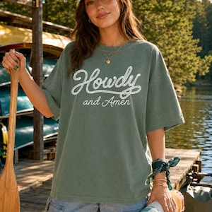 May include: An olive green short-sleeved t-shirt featuring the white text Howdy and Amen in a rope-style font. The shirt has a crew neckline and a relaxed fit. It is paired with light blue denim shorts and a beige baseball cap with text.