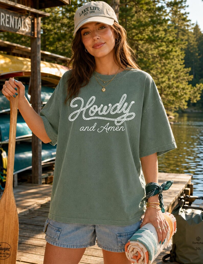 May include: An olive green short-sleeved t-shirt featuring the white text Howdy and Amen in a rope-style font. The shirt has a crew neckline and a relaxed fit. It is paired with light blue denim shorts and a beige baseball cap with text.