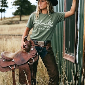 May include: An olive green t-shirt with the white text Howdy and Amen, a black cowboy hat, dark blue denim jeans, brown leather chaps with floral tooling, and a brown leather western saddle with silver hardware and intricate embossed patterns.