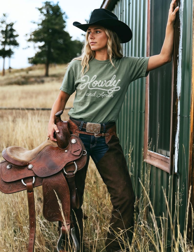 May include: An olive green t-shirt with the white text Howdy and Amen, a black cowboy hat, dark blue denim jeans, brown leather chaps with floral tooling, and a brown leather western saddle with silver hardware and intricate embossed patterns.