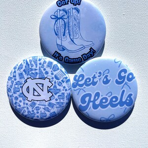 May include: Three light blue round buttons. One button has a drawing of cowboy boots and the text "Get Up!" and "It's Game Day!". Another button has the UNC logo and a pattern. The third button says "Let's Go Heels".