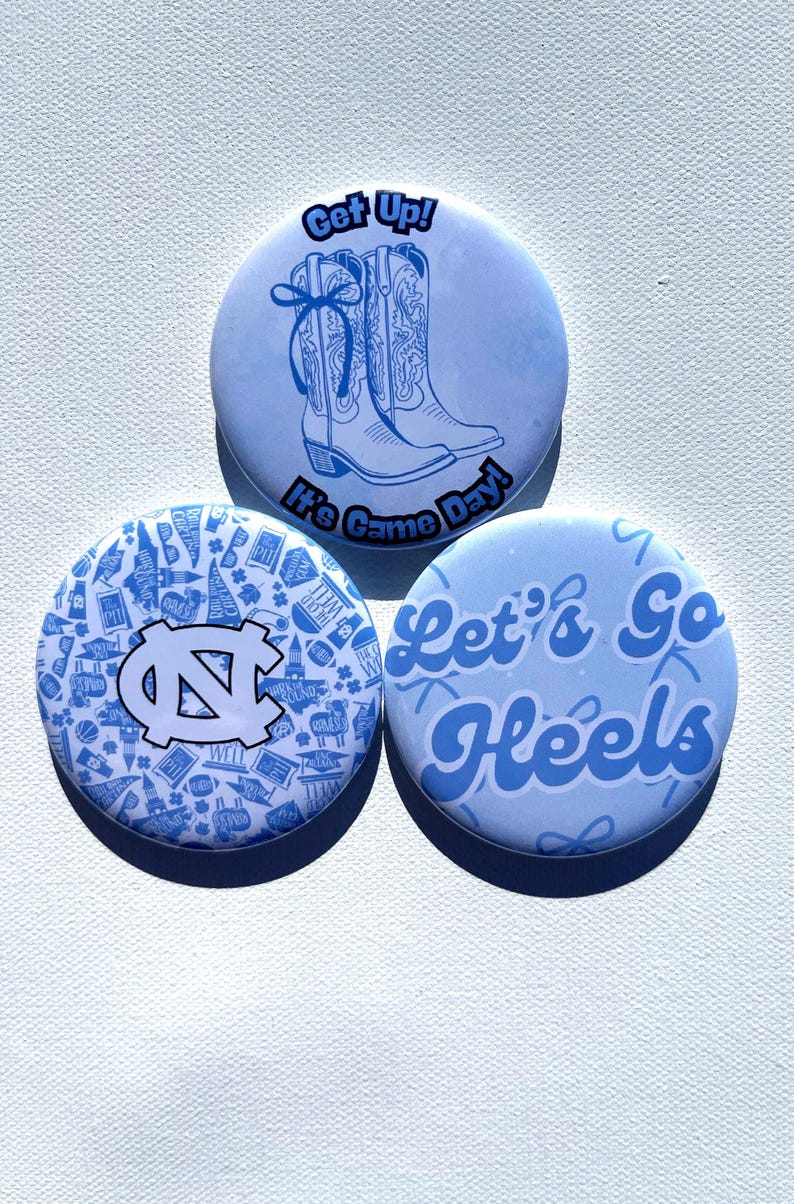 May include: Three light blue round buttons. One button has a drawing of cowboy boots and the text "Get Up!" and "It's Game Day!". Another button has the UNC logo and a pattern. The third button says "Let's Go Heels".