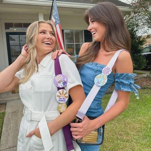 May include: Two women smiling, wearing white and denim outfits, each with a crossbody bag. One bag has a purple strap with decorative buttons that read "Tis the Season". The other bag has a white strap with a button that reads "Stau X".