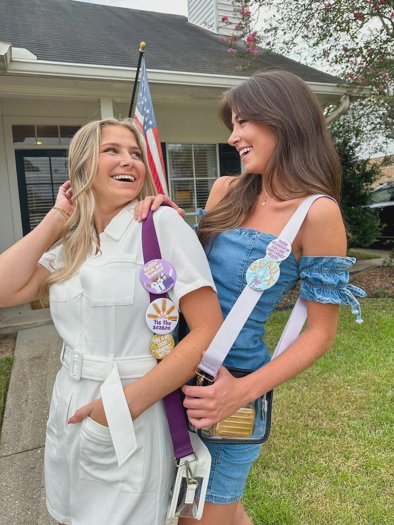 May include: Two women smiling, wearing white and denim outfits, each with a crossbody bag. One bag has a purple strap with decorative buttons that read "Tis the Season". The other bag has a white strap with a button that reads "Stau X".