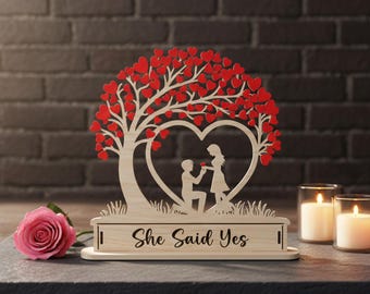Può includere: Scena di proposta tagliata al laser in legno. Un albero di cuori rossi, un ritaglio a forma di cuore con una figura in ginocchio e una donna. La base recita "She Said Yes". Una rosa rosa e candele sullo sfondo.