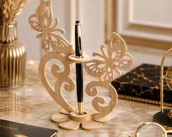 May include: A light-colored wooden pen holder featuring two intricate butterfly cutouts on either side of a central vertical pen slot. The base is shaped like a flower with rounded petals, and a black and gold pen is inserted into the holder.