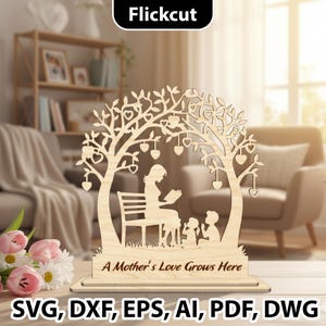 Puede incluir: Adorno decorativo de madera cortado con láser que representa a una madre leyendo a dos niños bajo un árbol con hojas en forma de corazón. El texto "A Mother's Love Grows Here" está en la base. La imagen también incluye el texto "Flickcut" y opciones de tipo de archivo.