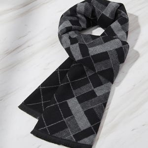 Puede incluir: Bufanda negra y gris con un patrón geométrico de rombos. La bufanda está hecha de un material suave y tiene un borde con flecos. La bufanda está doblada sobre una superficie de mármol blanco.