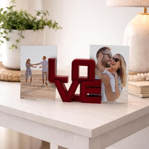 Könnte beinhalten: Roter Fotorahmen in Form des Wortes "LOVE". Er enthält zwei Fotos eines Paares. Der Rahmen steht auf einem weißen Tisch. Ein Blumentopf und eine Lampe sind im Hintergrund zu sehen. Dekoratives Objekt.