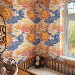 Op de afbeelding: Een kinderkamer met behang met een hemels ontwerp van lachende zonnen, manen en wolken in tinten geel, oranje, blauw en roze. Een houten wieg en een rieten mand met knuffels bevinden zich in de kamer.