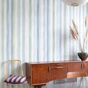 Op de afbeelding: Een kamer met een mid-century modern esthetiek. De muren zijn beschilderd met verticale strepen in tinten blauw, groen en wit. Een gouden stoel met een gestreept kussen staat naast een houten kast en een tafel met glazen blad.