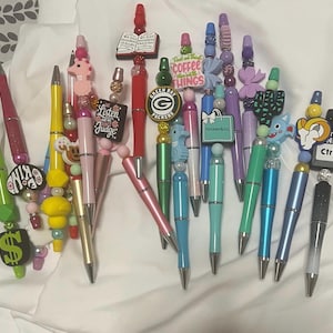 May include: A collection of decorative pens in various colors, featuring unique toppers. Some pens have charms with text like "Listen Judge" and "Coffee Things", and others have themed designs such as a seahorse, a cactus, and a Green Bay Packers logo.