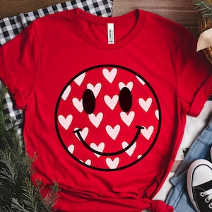 May include: A red t-shirt featuring a smiley face design filled with white hearts. The smiley face has black eyes and a black curved mouth. The shirt is made of a soft material and is paired with blue jeans and black sneakers.