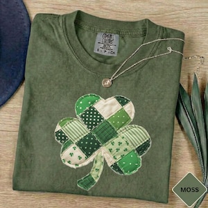 May include: A moss green Comfort Colors t-shirt with a patchwork shamrock design in various shades of green and white. A silver necklace with a pendant rests on the shirt. The shirt has a tag that reads "Comfort Colors".