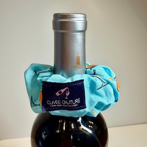 May include: A dark wine bottle with a silver cap and a light blue scrunchie. The scrunchie has a pattern of orange and black cocktail glasses. A navy blue label reads "CUVEÉ COUTURE" and "EVERY DROP TELLS A STORY".
