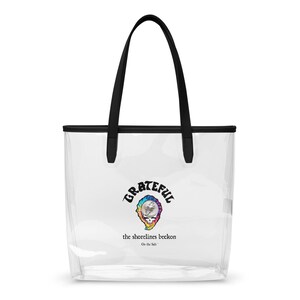May include: Clear tote bag with black trim and handles. The bag features a colorful graphic of a skull with the word "Grateful" above and the text "the shorelines beckon" below. The bag is designed for carrying items.