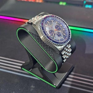 Peut inclure: Une montre-bracelet en acier inoxydable avec un cadran bleu et argenté, comprenant plusieurs sous-cadrans et un affichage de la date. La montre est présentée sur un support de montre noir avec une bordure verte. Le bracelet est également en acier inoxydable.