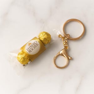 Peut inclure: Un porte-clés doré avec un mousqueton et un anneau rond. À côté du porte-clés se trouve un bonbon emballé dans du papier doré avec l'inscription "FERRERO ROCHER" sur l'étiquette.