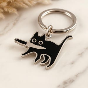 Könnte beinhalten: Ein schwarzer Katzen-Schlüsselanhänger mit einem silberfarbenen Metallring. Die Katze hat ein Messer im Maul und einen silbernen Umriss. Ein lustiges Accessoire.