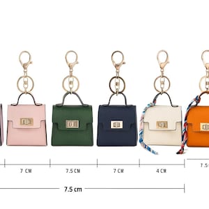 Könnte beinhalten: Eine Auswahl an Miniatur-Handtaschen-Schlüsselanhängern in den Farben Lavendel, Rosa, Grün, Marineblau, Weiß und Orange. Jede Tasche hat einen goldfarbenen Verschluss und Henkel sowie einen dekorativen Schal. Die Taschen sind ca. 7 cm breit und 7 cm hoch.