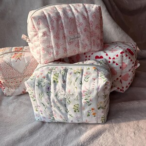 Peut inclure: Plusieurs trousses de maquillage matelassées avec différents motifs. L'une présente des nœuds roses sur fond blanc, une autre un imprimé floral avec le texte "Jengi Made". Chaque trousse a une fermeture éclair.