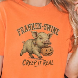 May include: An orange short-sleeve t-shirt featuring a graphic of a pig with stitches, bolts, and a jack-o'-lantern bucket. The text above the pig reads "FRANKEN-SWINE" and the text below reads "CREEP IT REAL" in a dark green, distressed font.