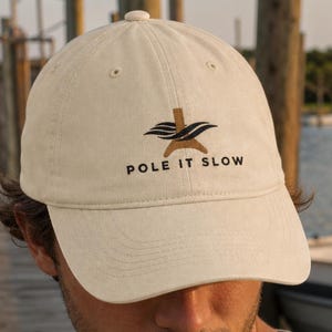 May include: An off-white baseball cap featuring a brown graphic of a stylized pole and black text that reads POLE IT SLOW. The cap has a structured crown, a curved brim, and a button on top, providing a clean and simple look for casual wear.