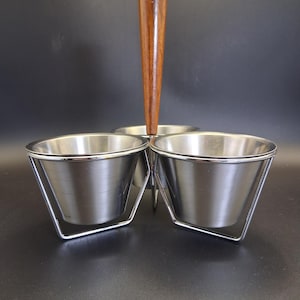 May include: A three-part serving dish featuring stainless steel bowls held in individual wire frames. The bowls are arranged around a central vertical post topped with a tall, tapered wooden handle for carrying the unit with ease.