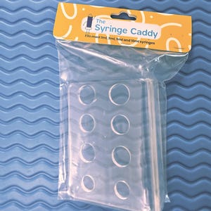May include: Clear plastic syringe caddy with eight circular openings, packaged in a clear plastic bag. The caddy has a yellow and white label that reads "The Syringe Caddy" and "Fits most 1ml, 3ml, 5ml and 10ml syringes."