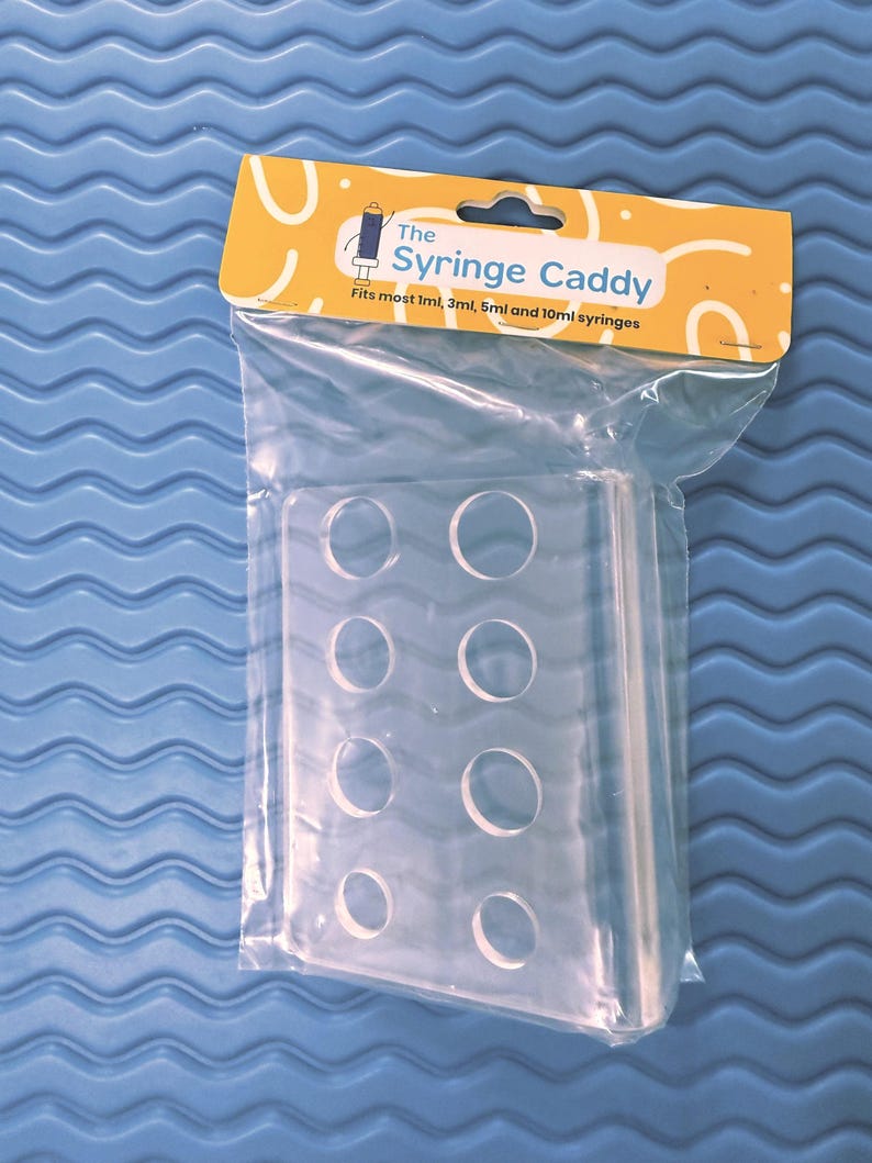 May include: Clear plastic syringe caddy with eight circular openings, packaged in a clear plastic bag. The caddy has a yellow and white label that reads "The Syringe Caddy" and "Fits most 1ml, 3ml, 5ml and 10ml syringes."