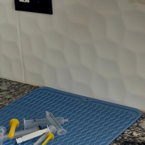 May include: A collection of syringes with yellow and white plungers rests on a blue silicone mat. A clear plastic holder for syringes is mounted on the wall. A black electrical outlet and light switch are visible.