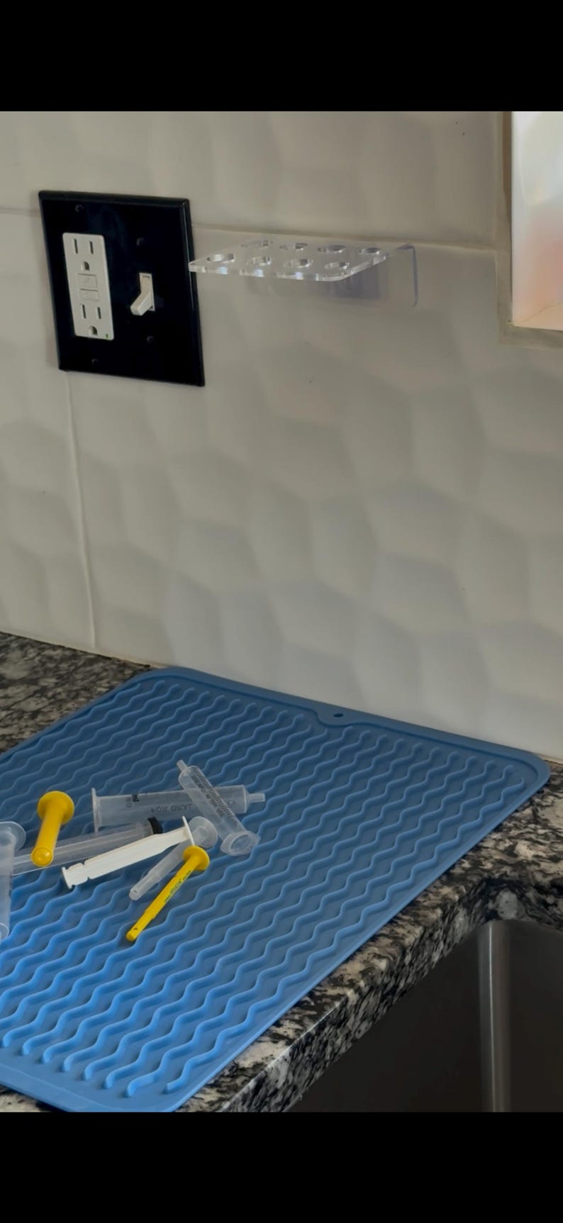 May include: A collection of syringes with yellow and white plungers rests on a blue silicone mat. A clear plastic holder for syringes is mounted on the wall. A black electrical outlet and light switch are visible.