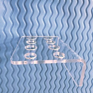 May include: A clear acrylic display stand with multiple circular openings. The stand has a rectangular top and a right-angled support leg. The background is a blue, wavy textured surface.