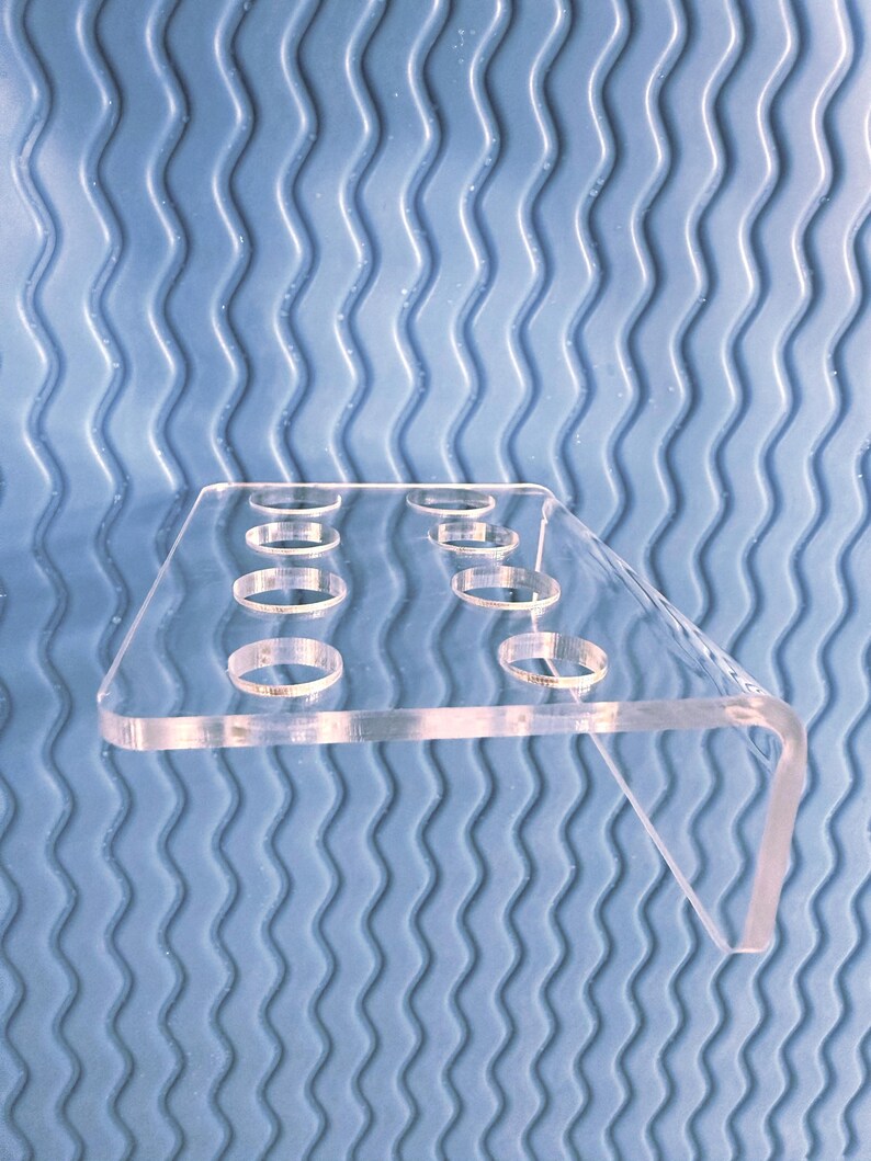 May include: A clear acrylic display stand with multiple circular openings. The stand has a rectangular top and a right-angled support leg. The background is a blue, wavy textured surface.