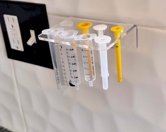 May include: A clear acrylic syringe holder with several syringes and plungers. The syringes are marked with measurements in milliliters. Some plungers are yellow, white, and clear. The holder is mounted on a white tiled wall.