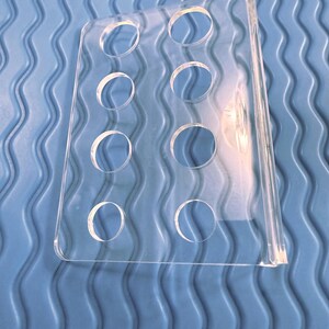 May include: A clear acrylic display stand with eight circular openings. The stand is rectangular with a curved top and a flat base. The background is a blue, wavy textured surface.