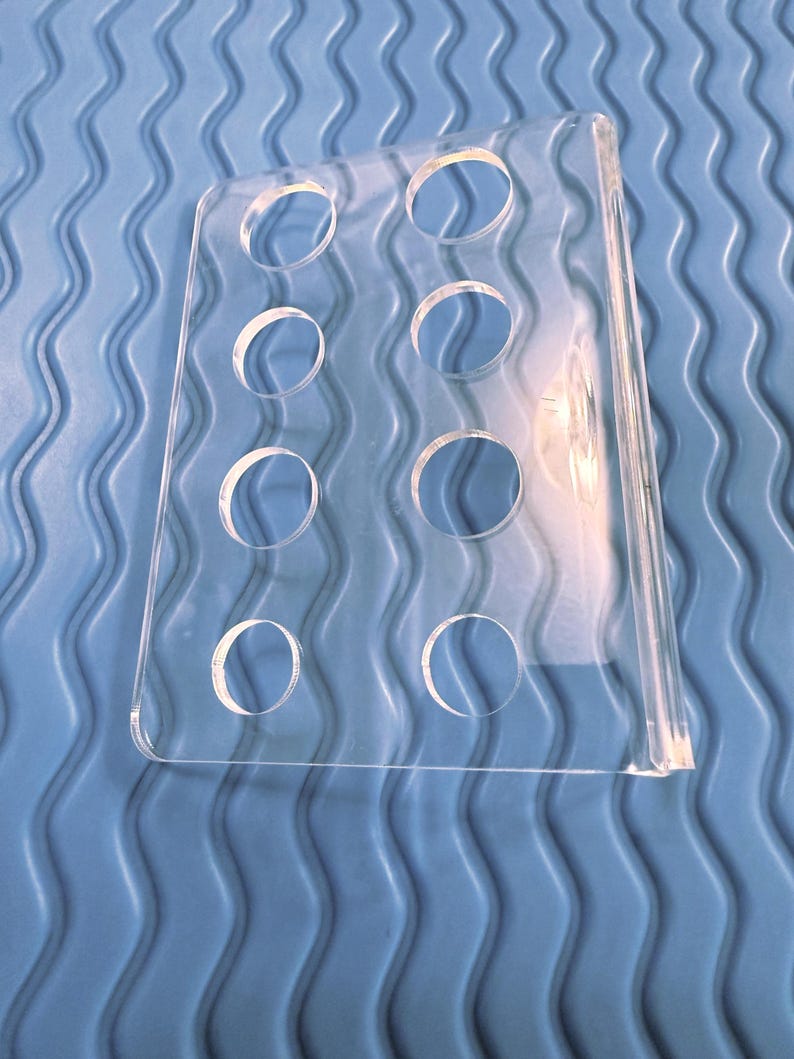 May include: A clear acrylic display stand with eight circular openings. The stand is rectangular with a curved top and a flat base. The background is a blue, wavy textured surface.