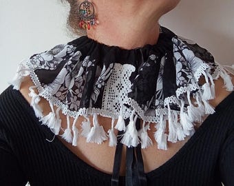 May include: A black and white floral patterned collar with white lace trim and white tassels. The collar is made of a lightweight fabric and has black satin ribbon ties at the back. A unique fashion accessory.
