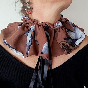 May include: A brown ruffled collar with a bird and branch pattern, tied with black ribbons. The fabric has a repeating design of white and blue birds. The collar is worn over a black top.