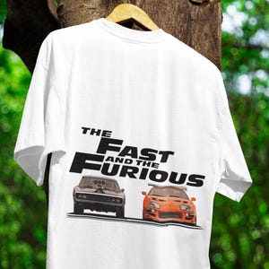 Puede incluir: Camiseta blanca con el texto "The Fast and the Furious" y ilustraciones de dos coches. Un coche es marrón oscuro, el otro es naranja. La camiseta está colgada en una percha de madera.