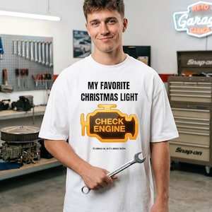 Pode incluir: Camiseta branca com o texto "MY FAVORITE CHRISTMAS LIGHT" acima de um gráfico da luz "CHECK ENGINE". Uma pessoa segura uma chave inglesa. O texto "It's always on, so it's always festive" está abaixo. A imagem é num garagem.