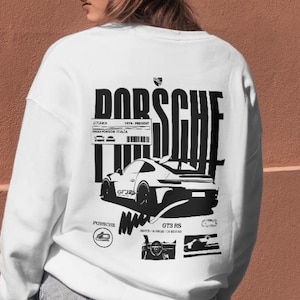 Pode incluir: Camisola branca com um gráfico preto de um carro desportivo e a palavra "PORSCHE" em negrito. O design inclui texto e ilustrações adicionais. A pessoa está a usar um boné de beisebol bege e calções de ganga.