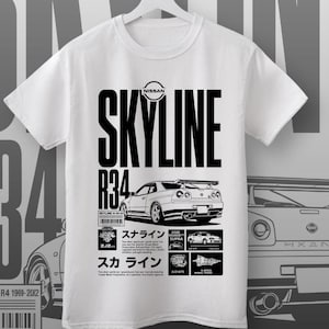 Puede incluir: Camiseta blanca con un gráfico negro de un coche Nissan Skyline R34. El diseño incluye el nombre del coche, detalles del modelo y texto japonés. La camiseta está colgada de una percha blanca sobre un fondo gris.