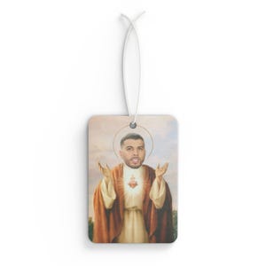 Può includere: Deodorante per ambienti rettangolare con un ritratto di un uomo in stile icona religiosa. L'immagine raffigura l'uomo con un'aureola, che indossa una veste e con un cuore luminoso. Un cordino bianco è attaccato per appenderlo.
