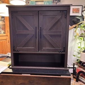 May include: A dark brown wooden cabinet with two doors and a lower shelf. The doors have a decorative X-shaped design. The cabinet has black hardware and is mounted on a larger wooden structure. The words "There's No Place Like Home" are visible above the cabinet.